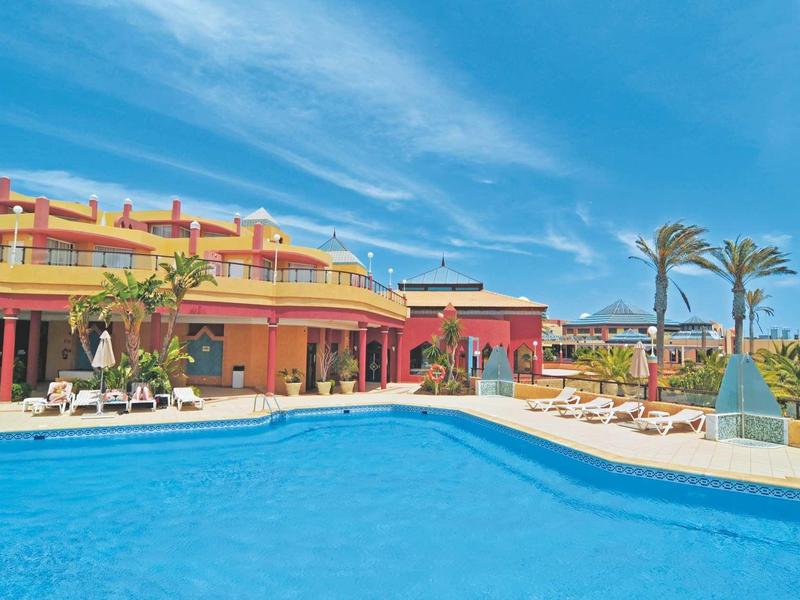 Grande piscina con lettini e complesso alberghiero tropicale sotto un cielo azzurro.