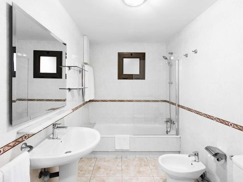 Weiß gefliertes Badezimmer mit Badewanne, Spiegel, Waschbecken und Toilette, schlicht gestaltet.