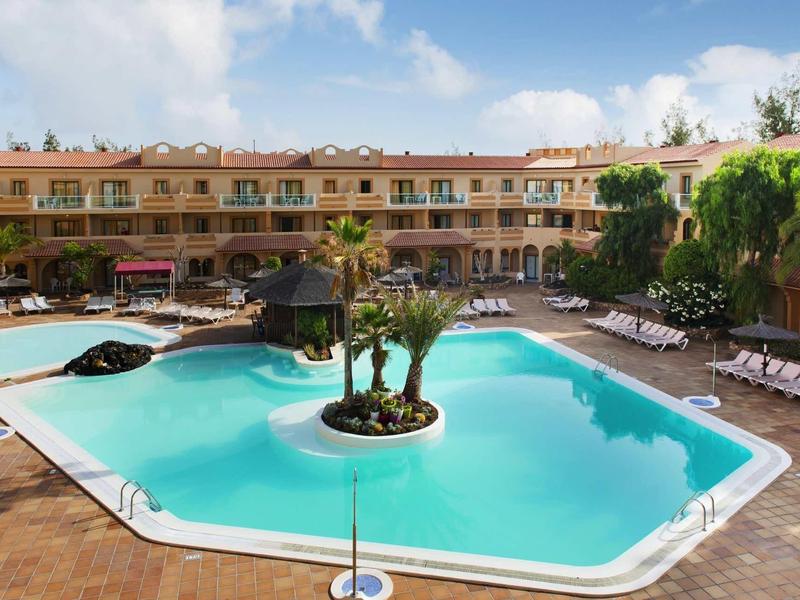 Schwimmbad mit türkisfarbenem Wasser, Sonnenliegen und Palmen in einem Innenhof mit zweistöckigen Gebäuden.