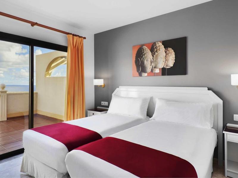 Modernes Hotelzimmer mit zwei weißen Betten, grauer Wand, rotem Bettläufer und Balkon mit Meerblick.