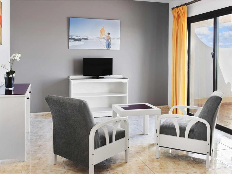 Moderner Wohnraum mit grauen Sesseln, weißem Tisch, Fernseher und orangenen Vorhängen.