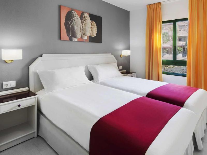 Modernes Hotelzimmer mit zwei weißen Betten, roten Deckenläufern, grauer Wand und orangen Vorhängen.