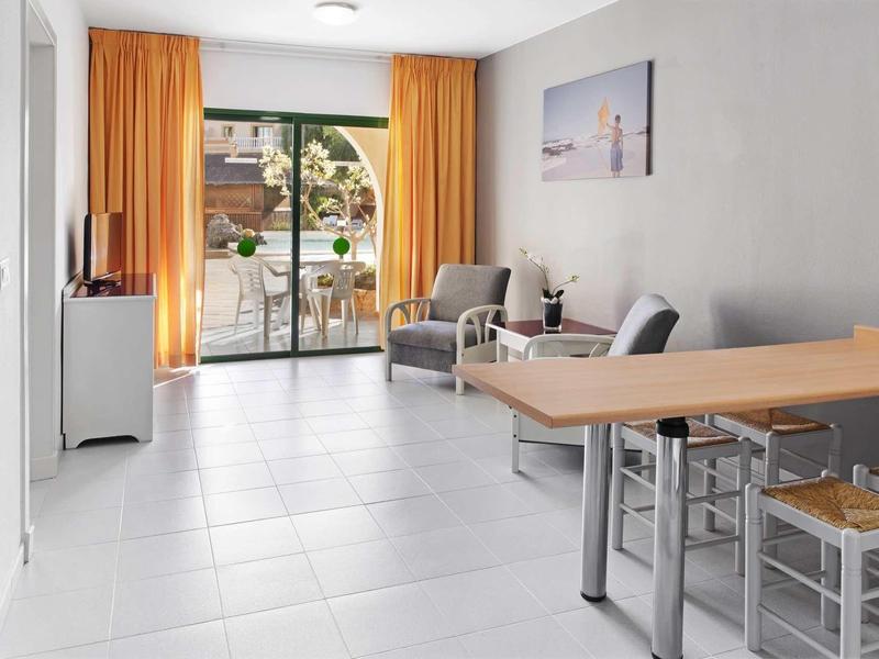 Heller Wohnraum mit weißem Boden, orangenen Vorhängen, grauen Sesseln und Holz-Esstisch am Fenster.