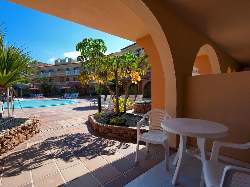 Sonnige Poolanlage mit blauen Liegestühlen, Pflanzen und Schatten unter Arkaden mit Kunststoffmöbeln.