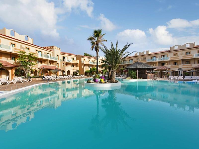 Großer Pool mit Palmen in der Mitte, umgeben von zweistöckigen Wohnungen unter blauem Himmel.