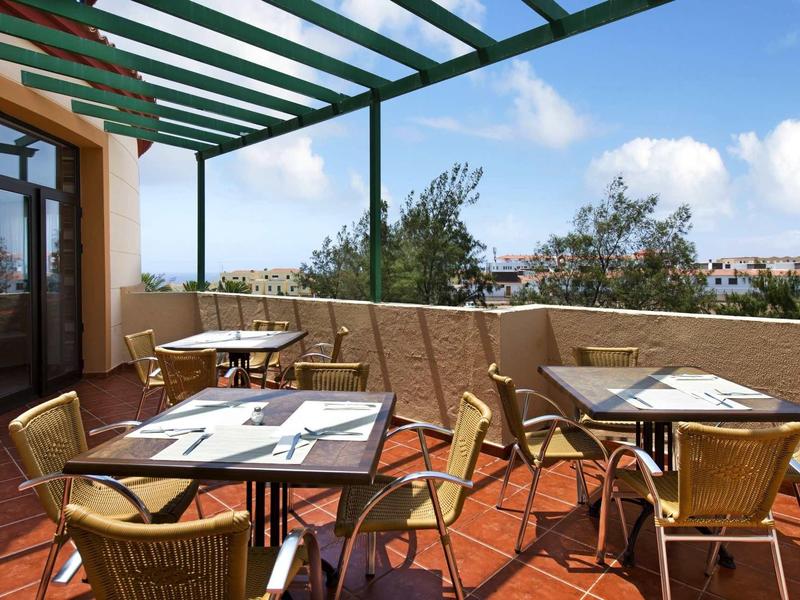 Dachterrasse mit Glasüberdachung, mehreren Tischen mit Stühlen und Blick auf Bäume und Himmel.