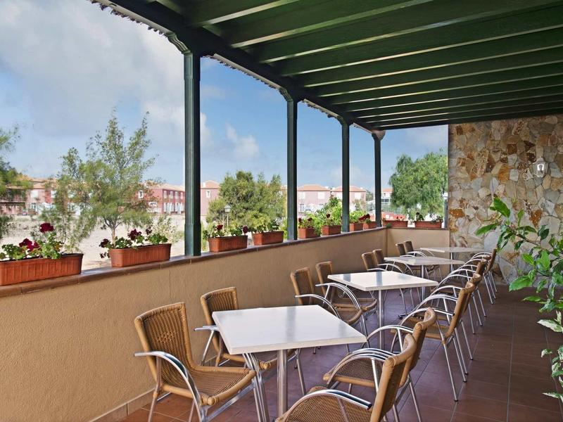 Überdachte Terrasse mit weißen Tischen, Metallstühlen und Pflanzen auf dem Geländer.