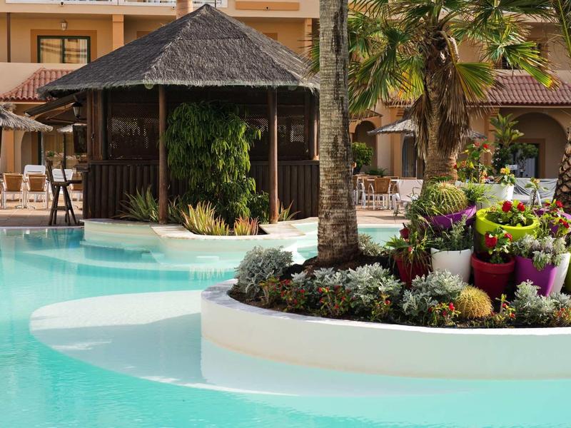 Großer Pool mit Palmeninsel und blühenden Pflanzen vor einem Holzpavillon und Gebäude im Hintergrund.