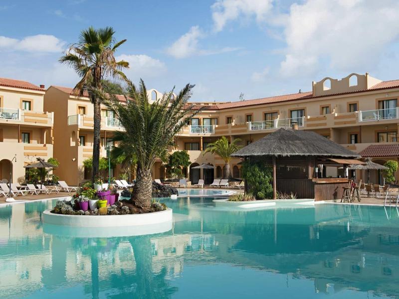 Großer Hotelpool mit Palmen und Liegestühlen vor einem gelben, mehrstöckigen Gebäude bei Sonnenschein.