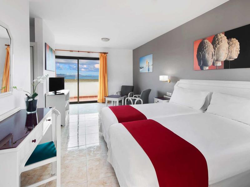 Helles Hotelzimmer mit Doppelbett, rotem Läufer, Schreibtisch, Fenster mit Meeresblick und grauer Wand.