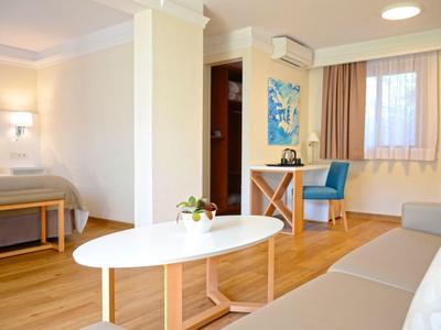 Helles Wohnzimmer mit Holzboden, Sofa, ovalem weißen Tisch, Schreibtisch mit Stuhl und Bett im Hintergrund.