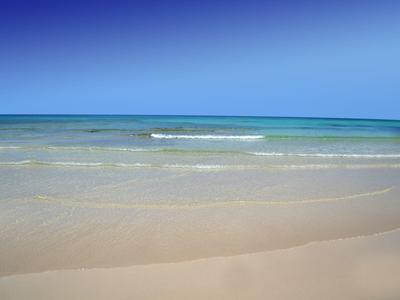 Widok na spokojną piaszczystą plażę z łagodnymi falami i czystym niebieskim niebem.