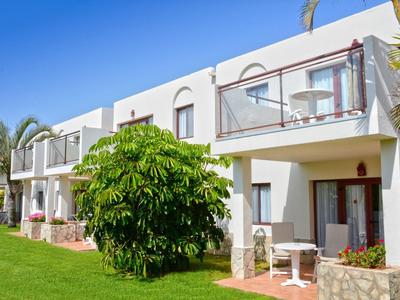 Weißes Reihenhaus mit Balkon, grünem Rasen und großem Busch bei klarem, blauem Himmel.