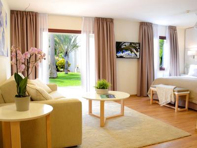 Heller Wohnraum mit Sofa, Couchtisch, Teppich, Bett und Vorhängen, Zugang zum Garten durch große Fenster.