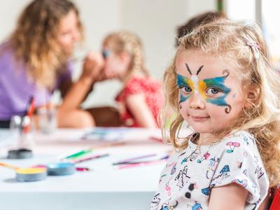 Ein Mädchen mit bunter Schmetterlings-Gesichtsbemalung sitzt im Vordergrund, im Hintergrund malt eine Frau.