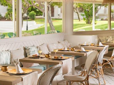 Helles Café mit Holztischen, weißen Stühlen und gepolsterten Sitzbänken nahe großen Fenstern.