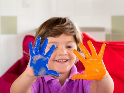 Ein lachender Junge zeigt seine Hände mit blauer und oranger Fingerfarbe vor sich.
