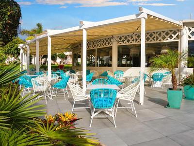 Terrasse mit weißen Möbeln, blauen Kissen und großen Pflanzen unter blauem Himmel.