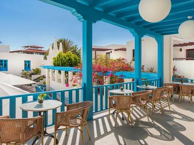 Balkon mit blauen Balken, runden Lampen, Korbstühlen und Tischen, Blick auf weiße Häuser und Pflanzen.