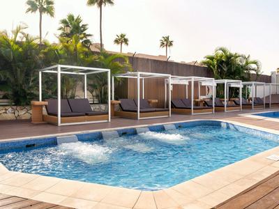 Pool mit blauer Wasserfläche, Umrandung aus Fliesen, vier weißen Badeliegen und Palmen im Hintergrund.