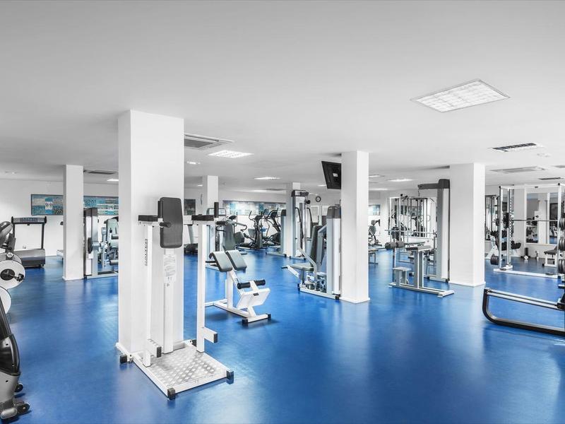 Moderner Fitnessraum mit blauem Boden, weißen Säulen, verschiedenen Trainingsgeräten und großen Fenstern.