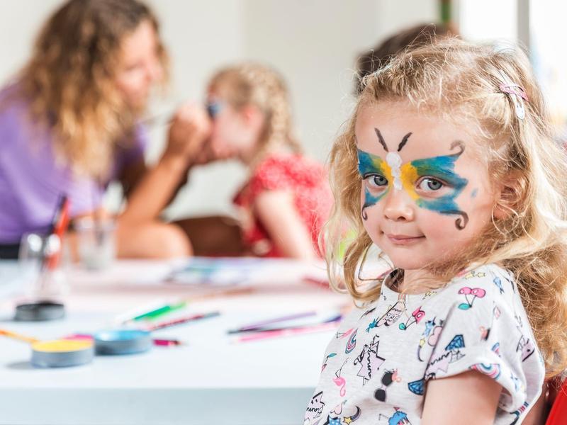Ein Mädchen mit bunter Schmetterlings-Gesichtsbemalung sitzt im Vordergrund, im Hintergrund malt eine Frau.