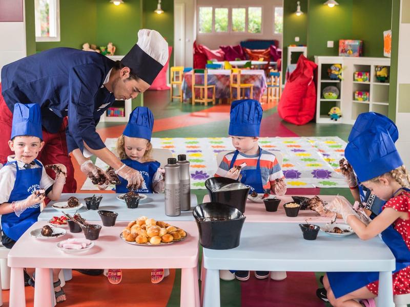 Kinder backen mit Kochmütze in buntem Raum, Lehrer hilft, Tische mit Gebäck und Utensilien.