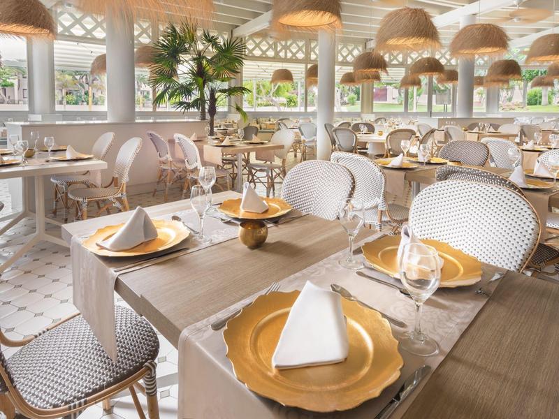 Eleganter Restaurant-Innenraum mit weißen Stühlen, Holztischen und goldfarbenen Tischdekorationen.