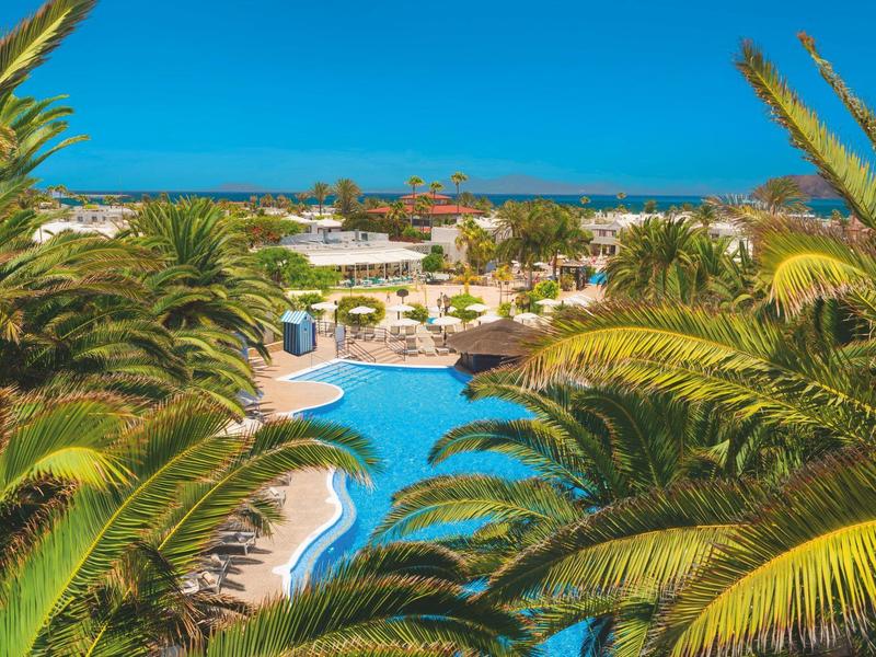 Ein blauer Pool umgeben von Palmen und Liegestühlen unter klarem blauem Himmel am Meer.