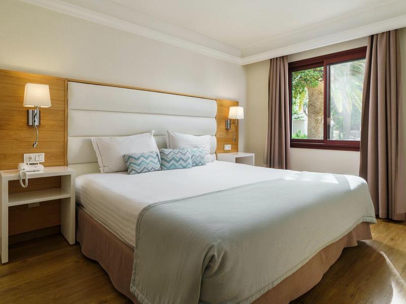 Helles Schlafzimmer mit großem Bett, Holzboden, Nachttischen, Wandlampen und Fenster mit Vorhängen.