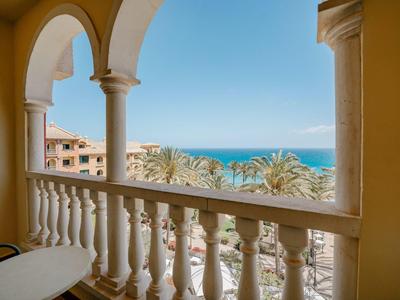 Balkon mit weißen Säulen, Blick auf Palmen, Gebäude und das blaue Meer unter klarem Himmel.