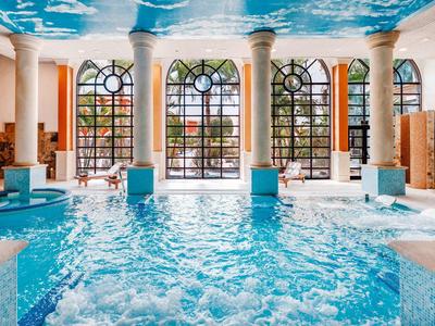 Ein heller, großer Indoor-Pool mit Säulen vor hohen Fensterbögen und einem Himmel-Deckenfresko.
