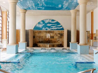 Helles Hallenbad mit Säulen, blauem Wasser und himmelblau bemalter Decke mit weißen Wolken.