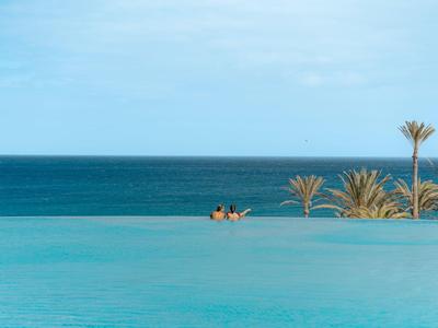 Ein endloser Pool mit blauem Wasser, dahinter das tiefblaue Meer und Palmen unter blauem Himmel.