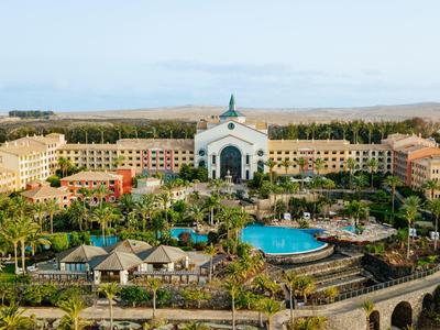 Großes Resort mit weißem Hauptgebäude, blauen Pool, Palmen und umliegender Landschaft.