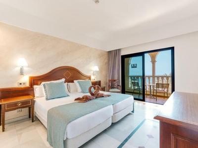 Helles Hotelzimmer mit zwei Einzelbetten, Holzmöbeln, Balkon und Meerblick bei Tageslicht.