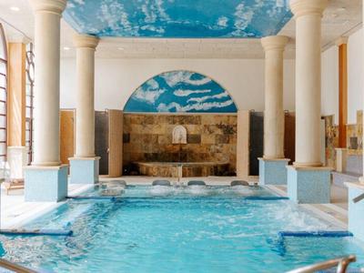 Hallenbad mit blauem Wasser, Marmorsäulen und einem Deckenfresko mit Wolkenmotiven.