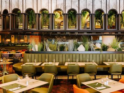 Modernes Restaurant mit grünen Sesseln, Holztischen, Pflanzen und dekorativen Spiegeln an der Wand.