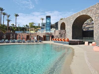 Piscine d'hôtel avec eau claire, arches en pierre et palmiers sous un ciel bleu.