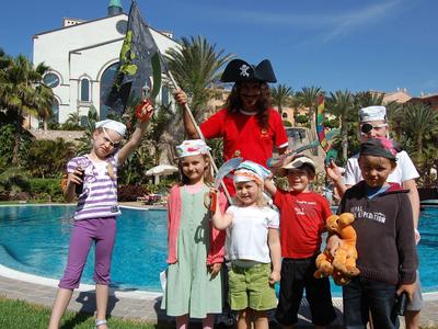Gruppe von Kindern und einem Erwachsenen in Piratenkostümen vor Pool und Gebäude bei Sonnenschein.