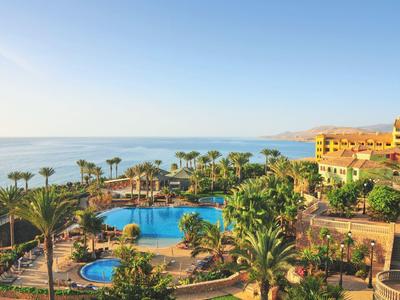 Hotelanlage mit mehreren Pools, Palmen und mediterranen Gebäuden am Meer bei klarem Himmel.