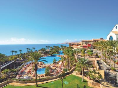 Blick auf Resort mit Pool, Palmen, Liegestühlen und Meer im Hintergrund unter blauem Himmel.