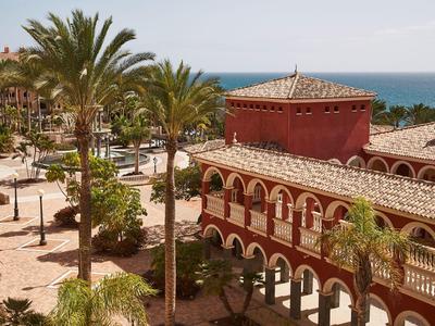 Rotes Gebäude mit Rundbögen, Palmen und Meer im Hintergrund an sonnigem Himmel.