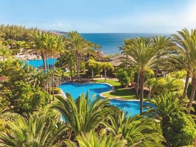 Tropische Gartenanlage mit Palmen, blauem Pool und Meer im Hintergrund bei klarem Himmel.