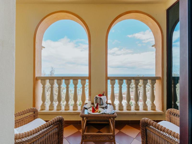 Balcon avec deux fauteuils en osier et une table avec des boissons, vue sur le ciel et la balustrade.
