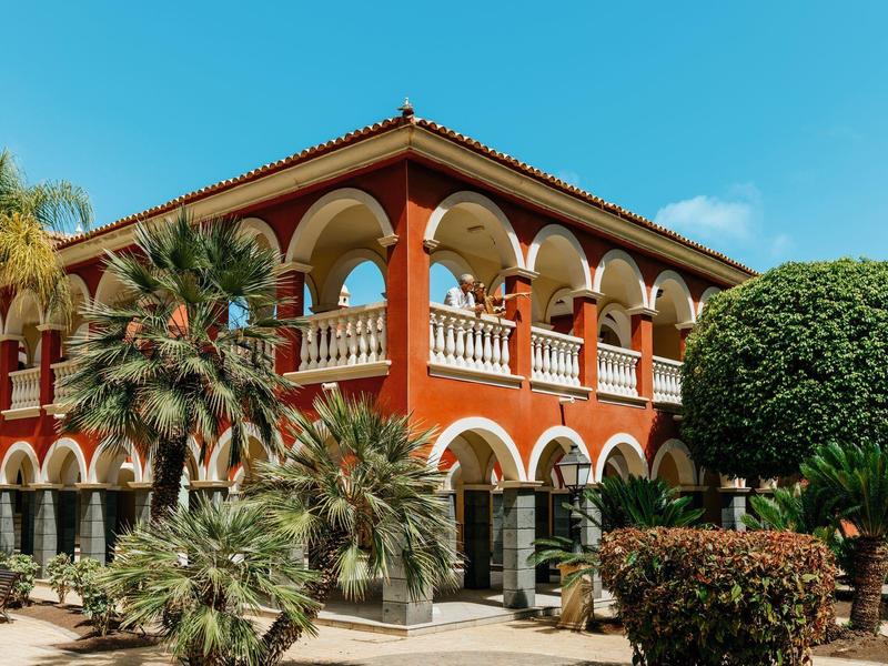 Hôtel aux murs rouges, arches et plantes tropicales sous un ciel dégagé.