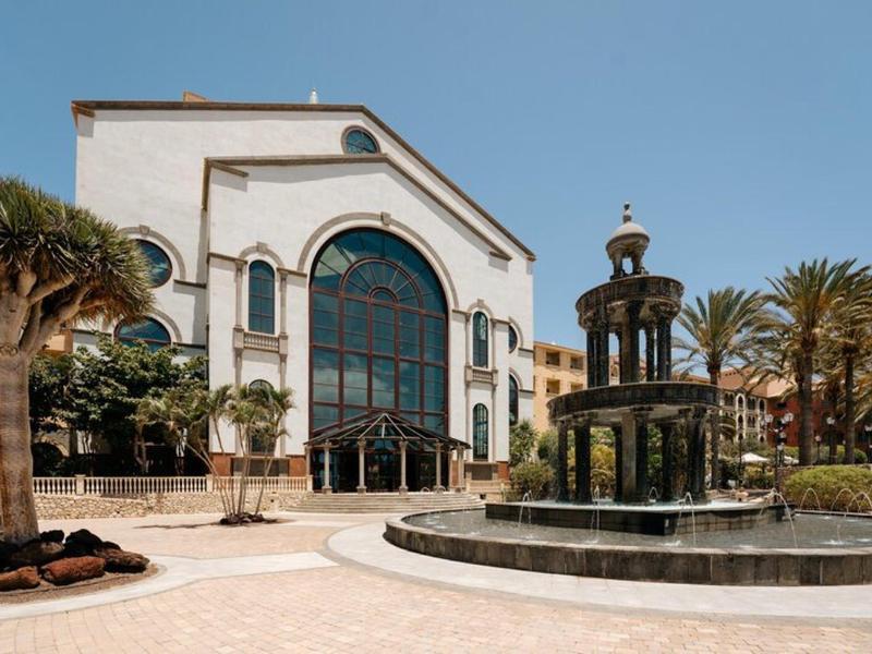Grand bâtiment d'hôtel blanc avec de grandes fenêtres et une fontaine devant, entouré de palmiers.