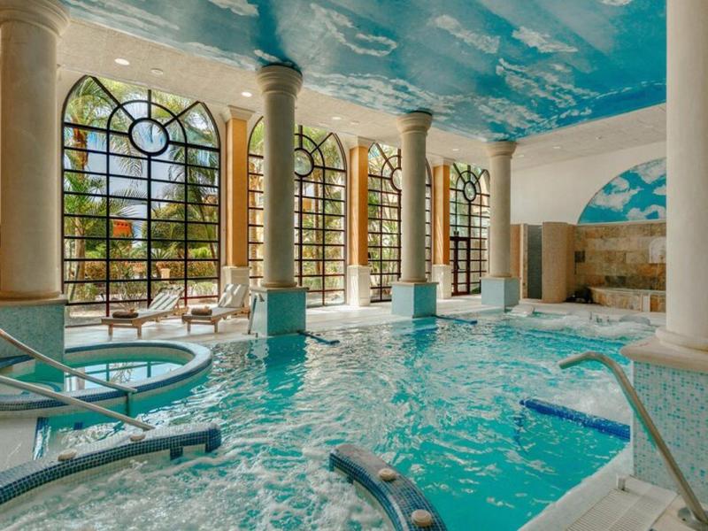 Piscine intérieure avec grandes fenêtres et colonnes sous un plafond décoratif représentant le ciel, jacuzzi à gauche.