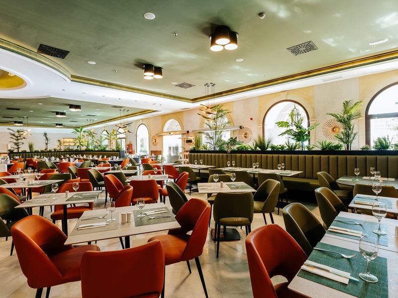 Restaurant lumineux et moderne avec chaises rouges, grandes fenêtres et tables dressées.
