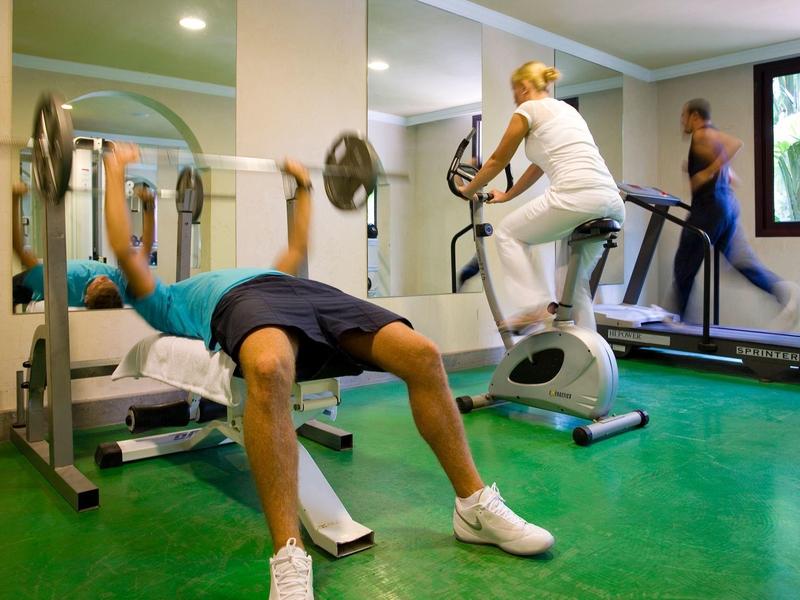 Trois personnes s'entraînent dans une salle de sport avec banc de musculation, vélo d'appartement et tapis de course.